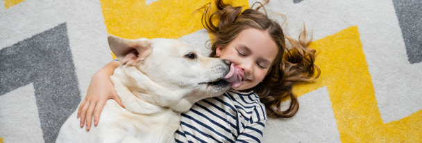 Horní pohled na labrador lízání obličeje pozitivního dítěte na podlaze doma, prapor  - Fotografie, Obrázek