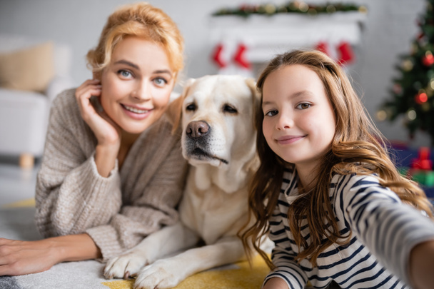 Smiling girl looking at camera near mom and labrador on floor at home  - Photo, Image