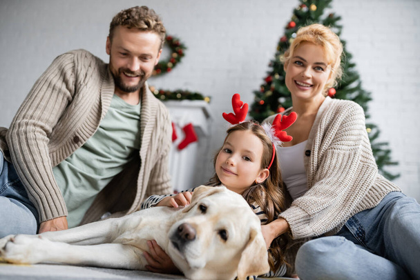 新年の間に家で床の上にぼやけたラブラドールの近くにカメラを見て幸せな家族  - 写真・画像