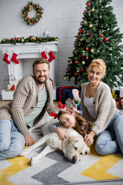 Famille positive avec fille et labrador assis près de l'arbre de Noël à la maison  - Photo, image