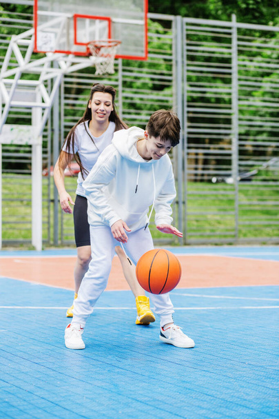 Girl and her younger brother, teenager, play basketball on modern basketball court under open sky. Concept of sports, hobbies and healthy lifestyle. - Foto, afbeelding
