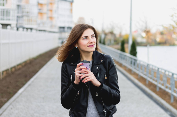 Menina bonita andando pela cidade com uma xícara de café, chá e sorrindo e desviando o olhar - Foto, Imagem