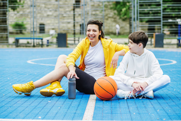 Teenager boy and girl are sitting on sports court, drinking water and resting during break. Basketball. Sport, leisure and family concept. - Photo, Image