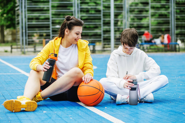 10代の男の子と女の子は、スポーツコートに座って、水を飲んで休憩中に休んでいます。バスケットボールだ。スポーツ、レジャー、家族のコンセプト. - 写真・画像
