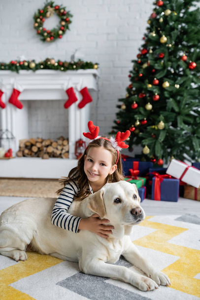 Cheerful girl petting labrador and looking at camera near christmas tree in living room  - Foto, imagen
