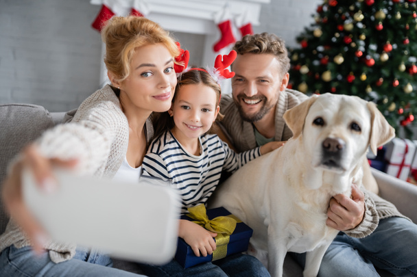 Женщина делает селфи с улыбающейся семьей и лабрадором во время Рождества дома  - Фото, изображение