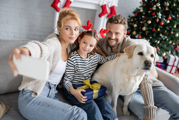 Uśmiechnięta rodzina z prezentem świątecznym i labradorem robiącym selfie na smartfonie w domu  - Zdjęcie, obraz
