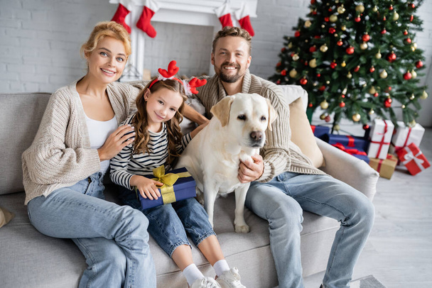 Uśmiechnięci rodzice przytulający córkę z prezentem świątecznym i labradorem w domu  - Zdjęcie, obraz