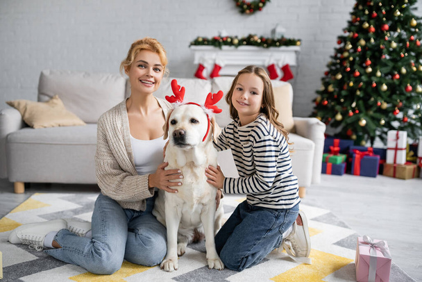 Smiling mother and daughter petting labrador with christmas headband near present at home  - Photo, Image