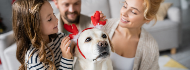 Meisje draagt nieuwe jaar hoofdband op labrador in de buurt wazig moeder en vader thuis, spandoek  - Foto, afbeelding