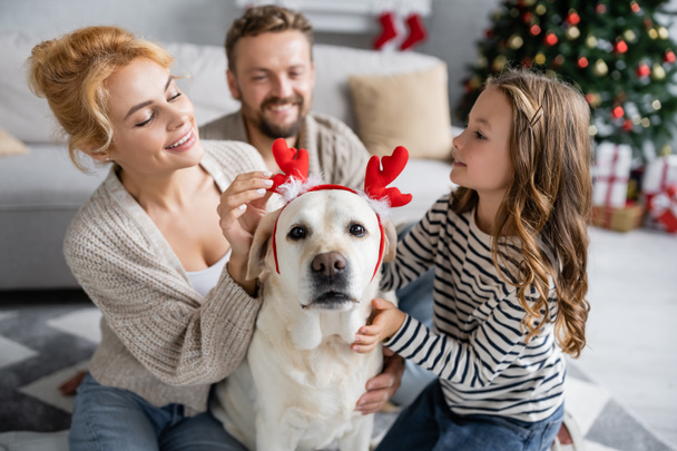 Mujer sonriente tocando diadema de Navidad en labrador cerca de la familia en la sala de estar  - Foto, Imagen