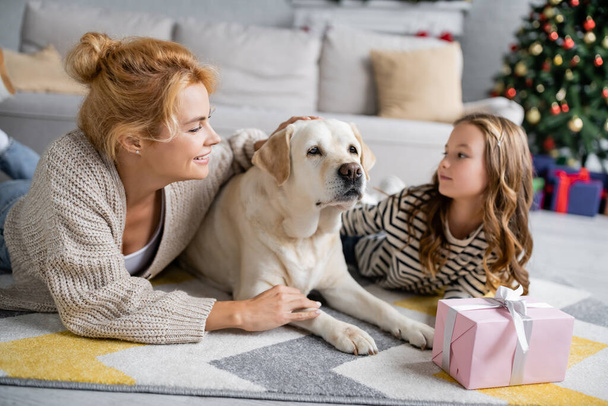 Femme gaie caressant labrador près de la fille et cadeau de Noël sur le sol à la maison  - Photo, image