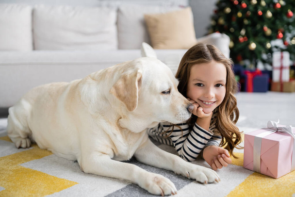 Labrador lying near smiling kid and christmas gift on carpet at home  - Фото, изображение