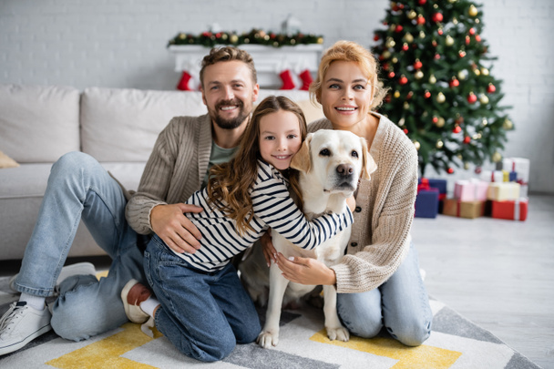 Щаслива сім'я з собакою-лабрадором дивиться на камеру під час Різдва вдома
  - Фото, зображення