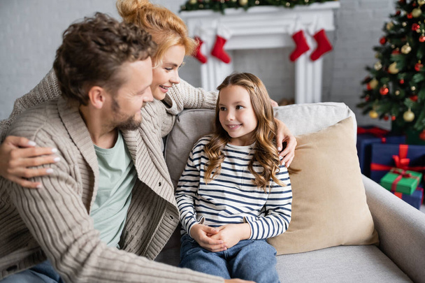 Mujer sonriente abrazando a su hija y esposo en el sofá durante la Navidad en casa  - Foto, Imagen