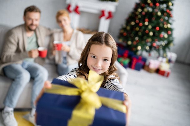 Fille gaie tenant cadeau de Noël et regardant la caméra près des parents flous dans le salon  - Photo, image