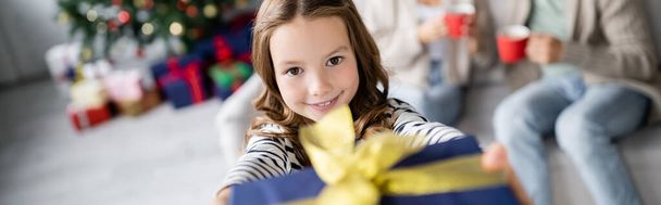 Smiling girl holding blurred christmas present and looking at camera at home, banner  - Foto, Imagen