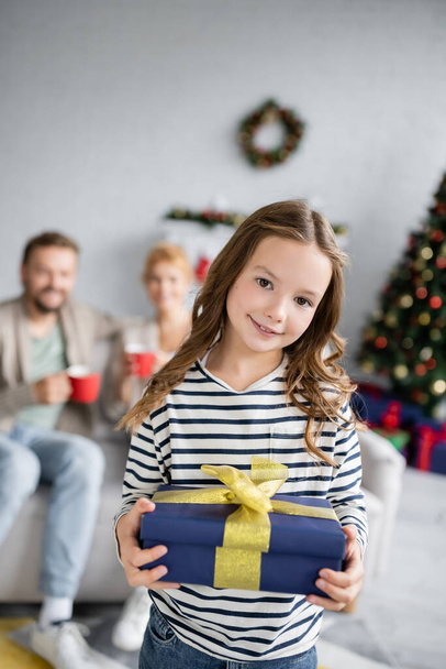 Kid holding new year gift near blurred family at home  - Zdjęcie, obraz