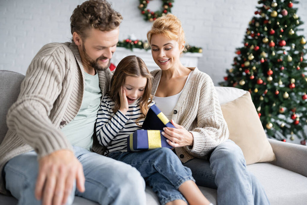 Ragazza eccitata guardando scatola regalo aperta vicino ai genitori durante il Natale a casa  - Foto, immagini