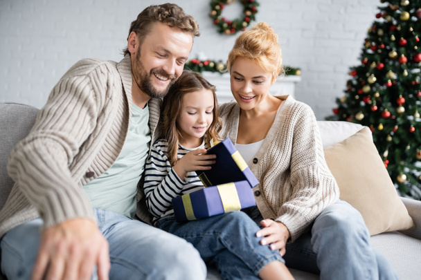 Des parents souriants regardant leur fille ouvrir un cadeau de Noël sur le canapé à la maison  - Photo, image