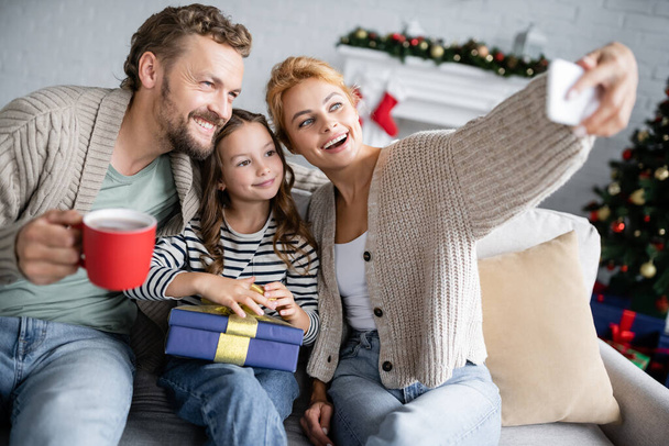 Frau macht Selfie mit Familie mit Weihnachtsgeschenk und Tasse auf der heimischen Couch  - Foto, Bild