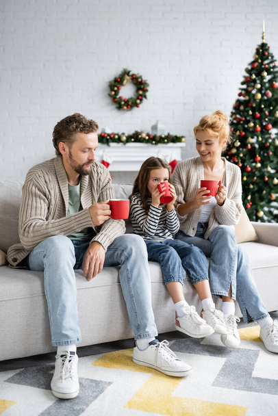 Девушка пьет какао рядом с родителями на диване во время Рождества дома  - Фото, изображение