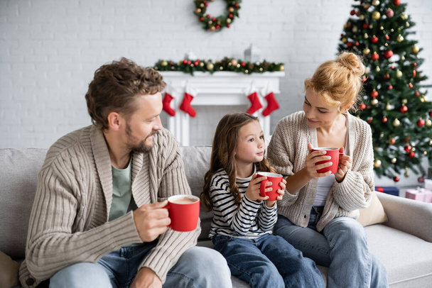 Улыбающиеся родители смотрят на дочь с какао на диване в новом году  - Фото, изображение