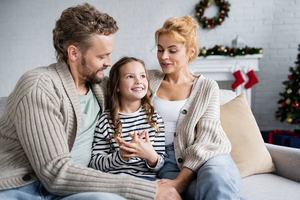 Zufriedenes Kind sitzt zu Weihnachten zu Hause neben Eltern auf der Couch  - Foto, Bild