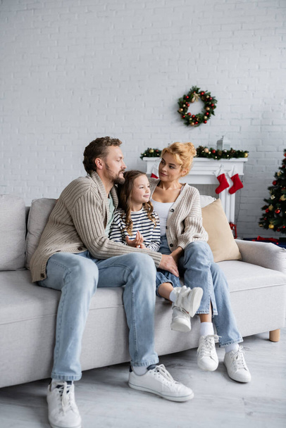 Smiling parents hugging daughter on couch near blurred christmas decor at home  - Photo, Image