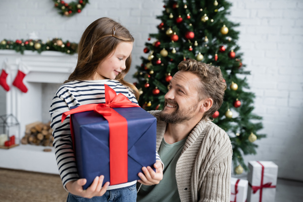 Smiling dad looking at daughter with present near blurred christmas tree at home  - Photo, Image