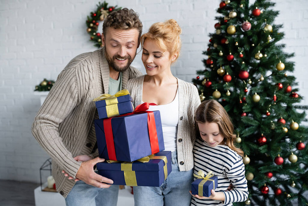 Smiling man and woman looking at gifts near daughter and christmas tree at home  - Фото, изображение