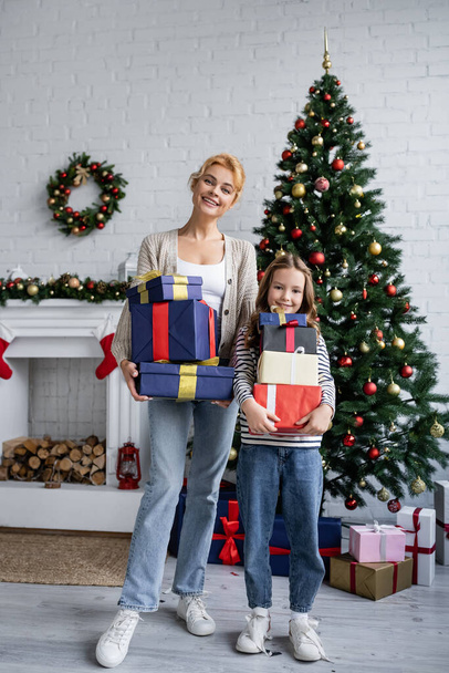 Cheerful mother and daughter holding presents near christmas tree and fireplace at home  - 写真・画像