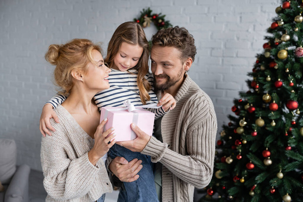Smiling man holding gift and daughter near wife and christmas tree at home  - Φωτογραφία, εικόνα