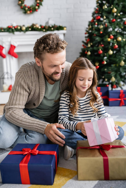 Smiling girl holding new year gift near dad on carpet in living room  - Photo, Image