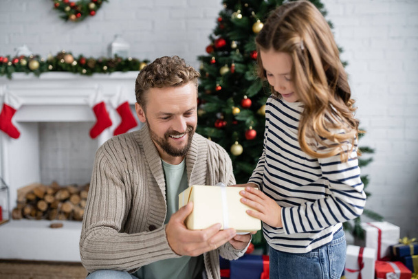 Homme positif en tricot cardigan tenant boîte cadeau près de l'enfant pendant Noël à la maison  - Photo, image