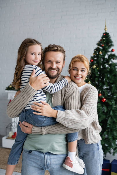 Positive man holding daughter near wife in cardigan and blurred christmas tree at home  - Foto, afbeelding