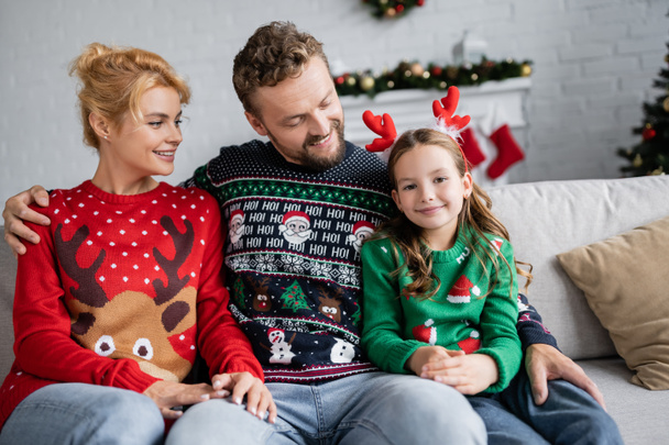 Homme joyeux en pull de Noël regardant l'enfant et étreignant femme sur le canapé à la maison  - Photo, image