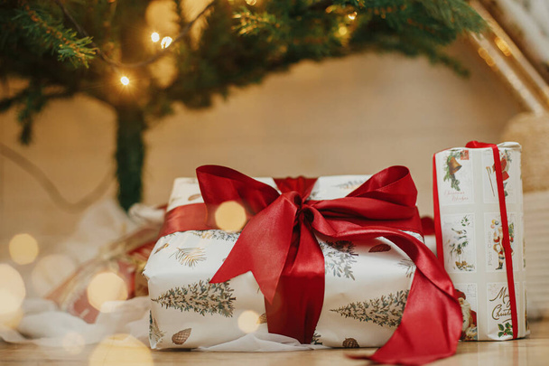Stijlvolle kerstcadeaus onder kerstboom met gouden lampjes bokeh. Verpakte kerstcadeaus met rood lint onder versierde boom met verlichting in de kamer. Atmosferische avond. Vakantiebanner - Foto, afbeelding