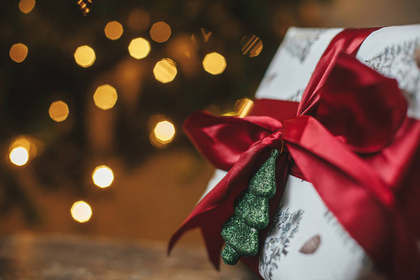Elegante regalo di Natale all'albero di Natale con luci dorate. Regalo di Natale avvolto con fiocco rosso e ornamento di albero lucente in camera di sera. Buon Natale! Stendardo atmosferico, spazio per il testo - Foto, immagini
