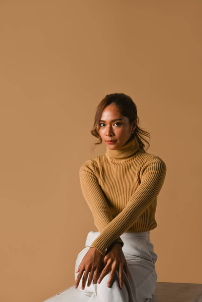 Foto de estudio de moda, concepto de otoño e invierno. Mujer joven atractiva en suéter de moda sentado sobre fondo de estudio marrón. - Foto, Imagen