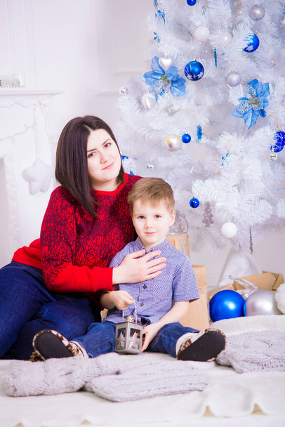 A young mother with dark hair with her son near a white Christmas tree  in a room decorated for Christmas. Family Christmas. Christmas mood - Valokuva, kuva