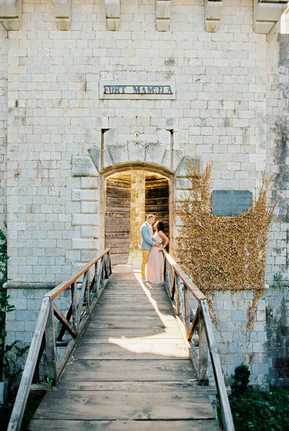 Groom hugs bride leaning against the railing of the wooden bridge to Mamula Fort. High quality photo - Photo, Image