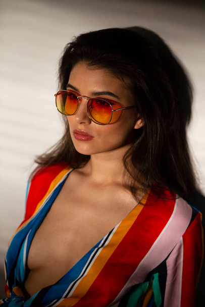 Portrait of a young seductive elegant woman in colorful top and sunglasses. Art portrait with shadows. Sunset light. - Foto, Bild