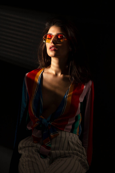 Εμπρός πορτρέτο μιας νεαρής αισθησιακής νεαρής γυναίκας με πολύχρωμα ρούχα και γυαλιά ηλίου, με κλειστά μάτια, σκούρο φόντο. Κάθετη προβολή. - Φωτογραφία, εικόνα