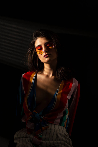 Front portrait of a young brunette young woman in colorful clothes and orange sunglasses, with closed eyes, dark background. Vertical view. - Foto, Bild