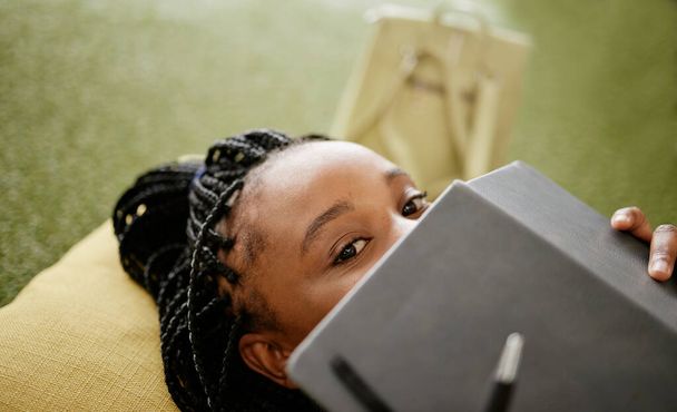 Schwarze Frau und Studentin verstecken Gesicht mit Notizbuch auf lässiger College-Lounge-Sitztasche. Schüchternes afrikanisches Universitätsstudium: Pause mit Hörbuch, um Identität zu verbergen - Foto, Bild