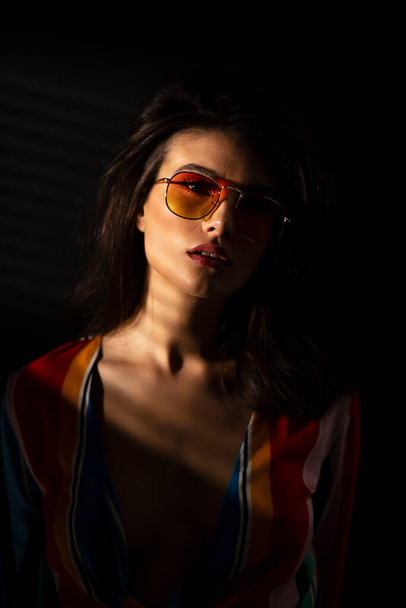 Front image of a young brunette woman in colorful clothes and orange sunglasses, dark background. - Fotografie, Obrázek
