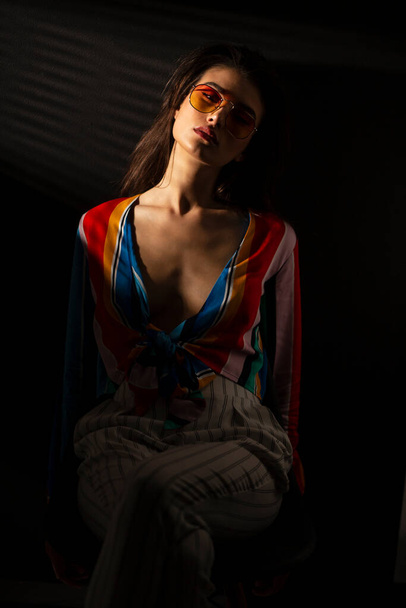 Передний портрет молодой брюнетки в разноцветной одежде и оранжевых солнцезащитных очках, сидящей на стуле, изолированный черный фон. - Фото, изображение