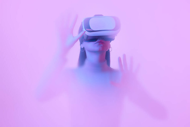 Virtual reality. Young girl in VR glasses, immersed in clouds of smoke, fog on a pink neon background with a backlight diffuser. Concept of art, metaverse, technology. Blurring effect - Zdjęcie, obraz