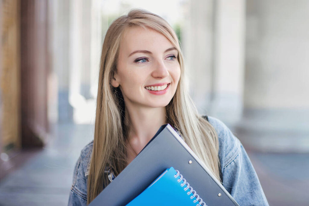 Close up retrato de uma menina branca sorridente estudante loira com olhos azuis em pé no fundo de uma antiga universidade e segurando pastas e cadernos - Foto, Imagem